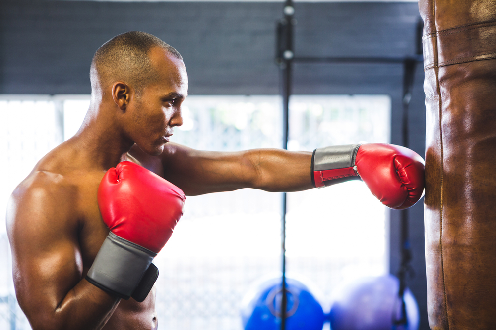 Side view of shirtless male boxer punching bag in fitness studio